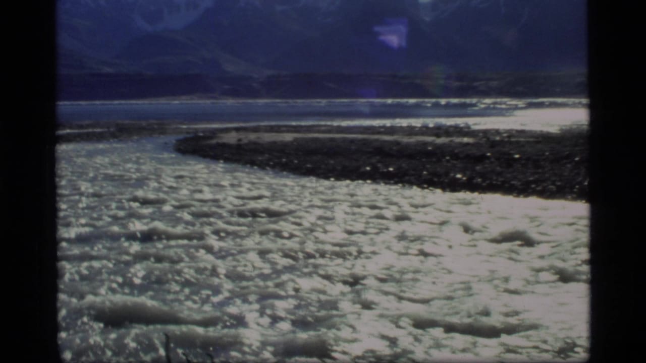 Rushing river with white foam, rocky shore, and distant mountains.