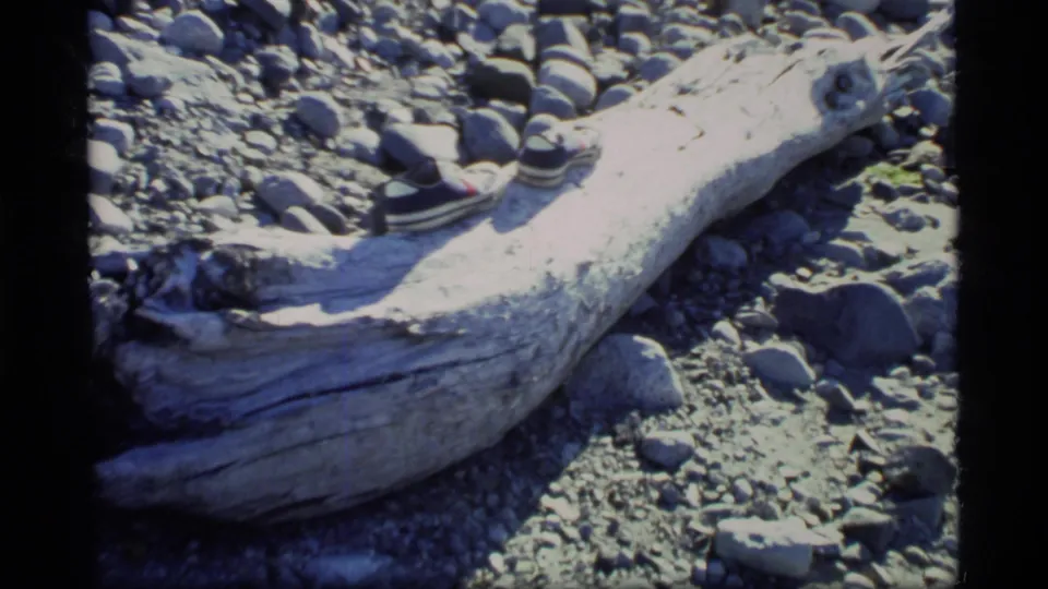 1977 Glacier Bay 8mm frame with a pale driftwood log, sneakers, and scattered stones in harsh sun near the end of the shot.