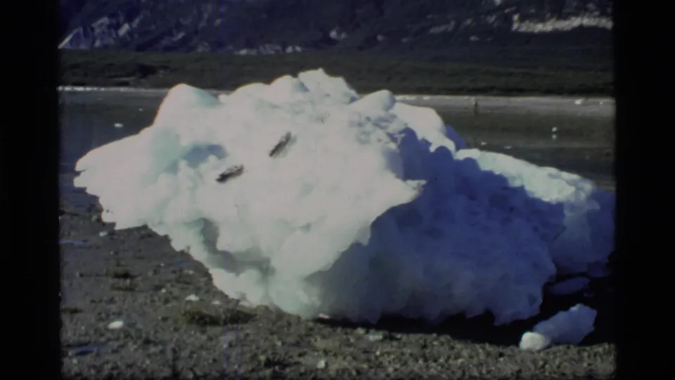 1977 Glacier Bay home-movie frame of a shoreline iceberg under strong light, backed by water and mountains.
