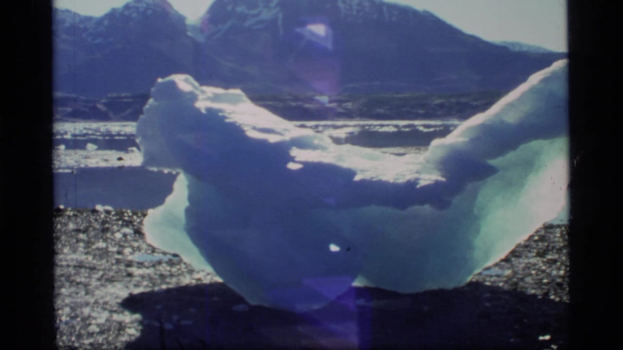 1977 Glacier Bay home-movie frame showing a sculpted iceberg near shore with dark water, snow-capped mountains, film borders, and lens flare.