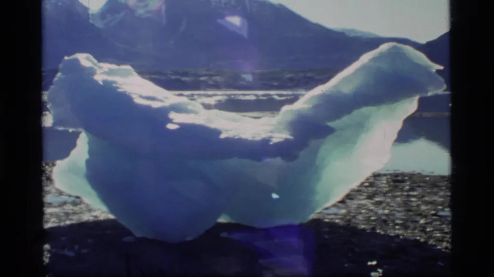 1977 Glacier Bay frame with a close view of a translucent iceberg, rippled water, distant mountain slopes, and visible 8mm film edge shadows.