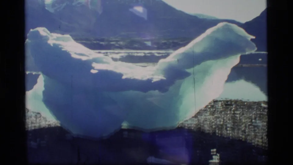 1977 Glacier Bay 8mm frame showing the iceberg from a shifted handheld angle, with blue-green ice, dark shoreline water, mountains, and film grain.