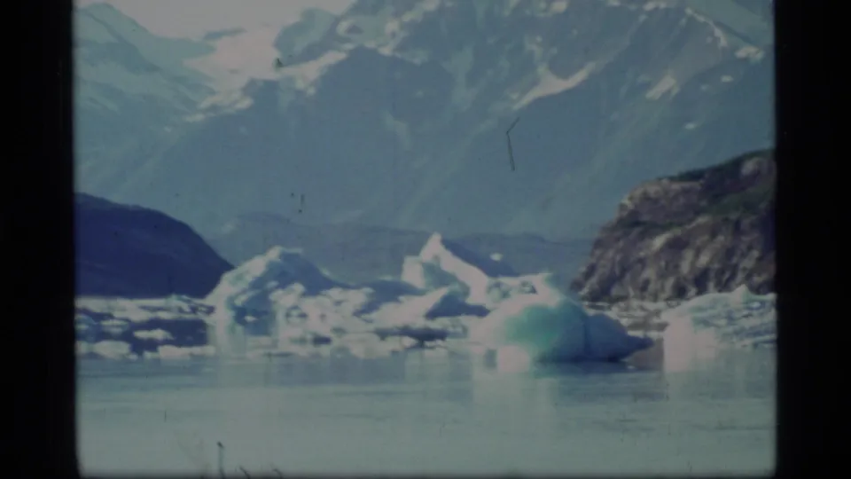 1977 Glacier Bay frame with smaller ice pieces on calm water beneath high snow-covered mountains and a rocky slope at the right edge.