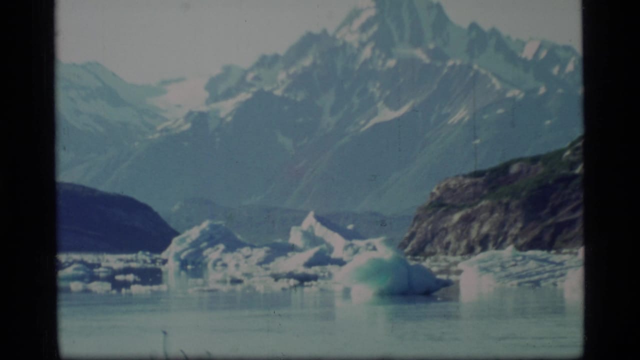 1977 8mm view of Glacier Bay with floating ice, calm water, distant mountains, and a dark rocky shore.