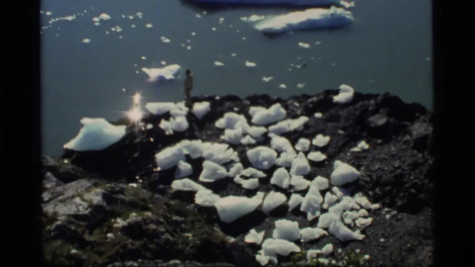 A lone distant figure stands on a rocky Glacier Bay shore amid ice chunks, with reflective water and floating ice beyond.