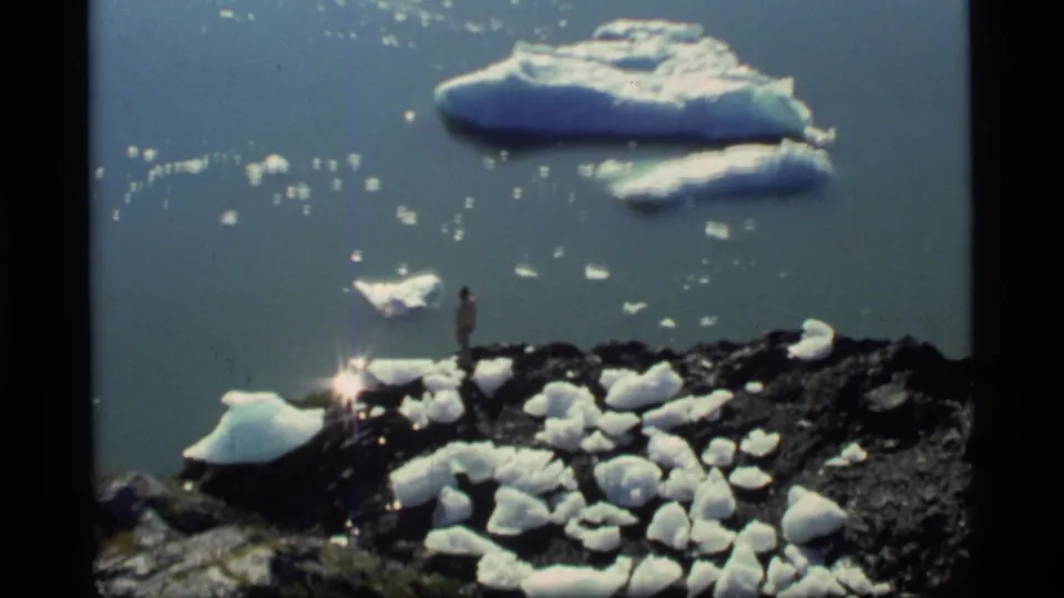 1977 Glacier Bay home-movie frame showing a distant person on rocky shore, foreground ice chunks, and larger icebergs offshore.