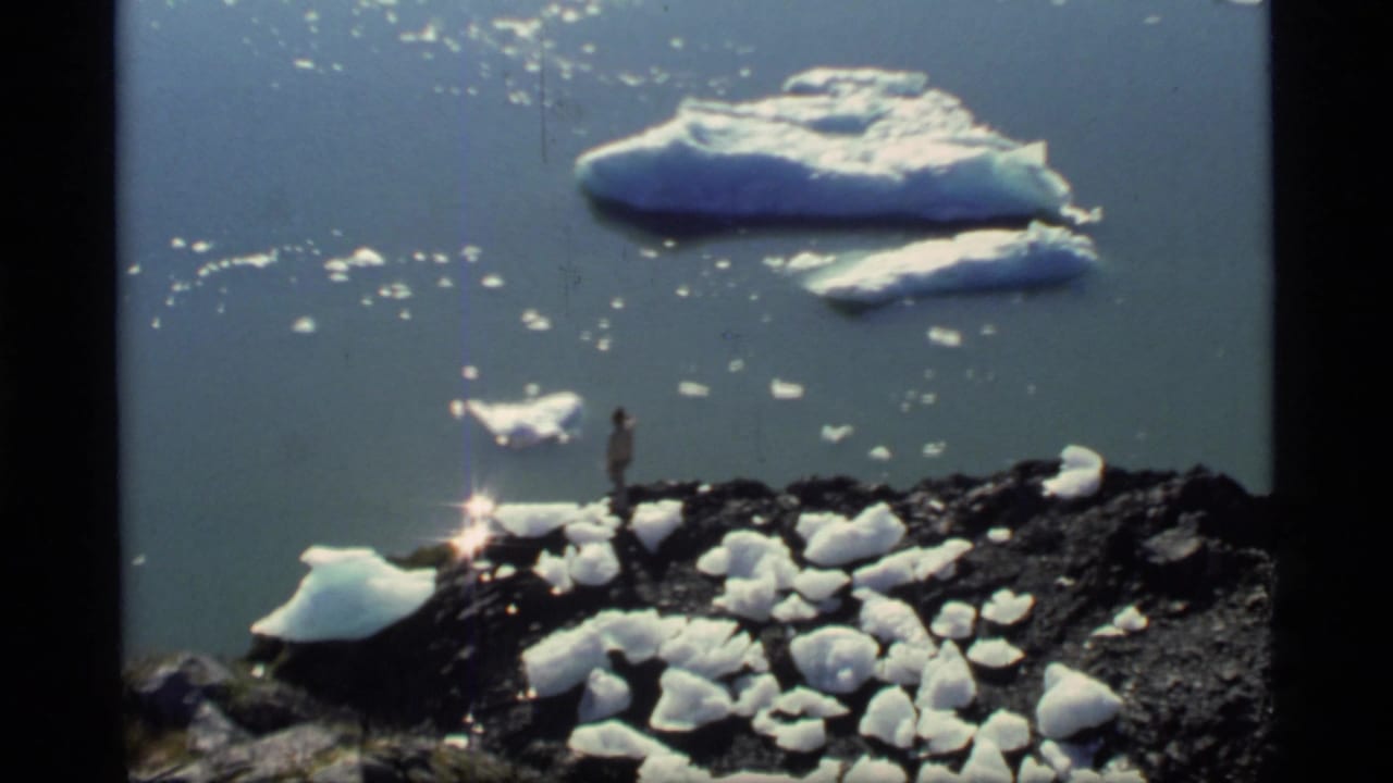 1977 Glacier Bay home-movie frame showing a small figure on a rocky, ice-dotted shoreline with sunlit water and floating ice beyond.