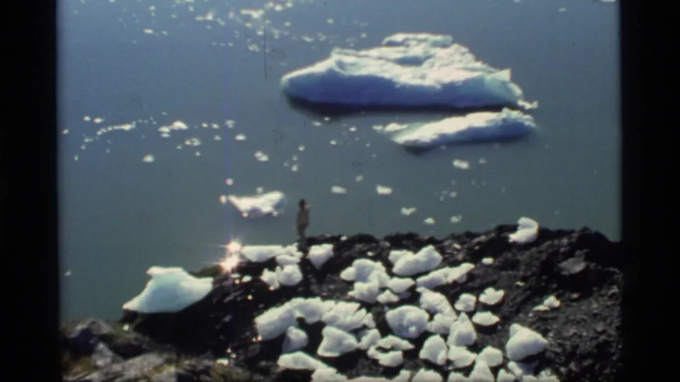 1977 Glacier Bay home-movie frame showing a small figure on a rocky, ice-dotted shoreline with sunlit water and floating ice beyond.