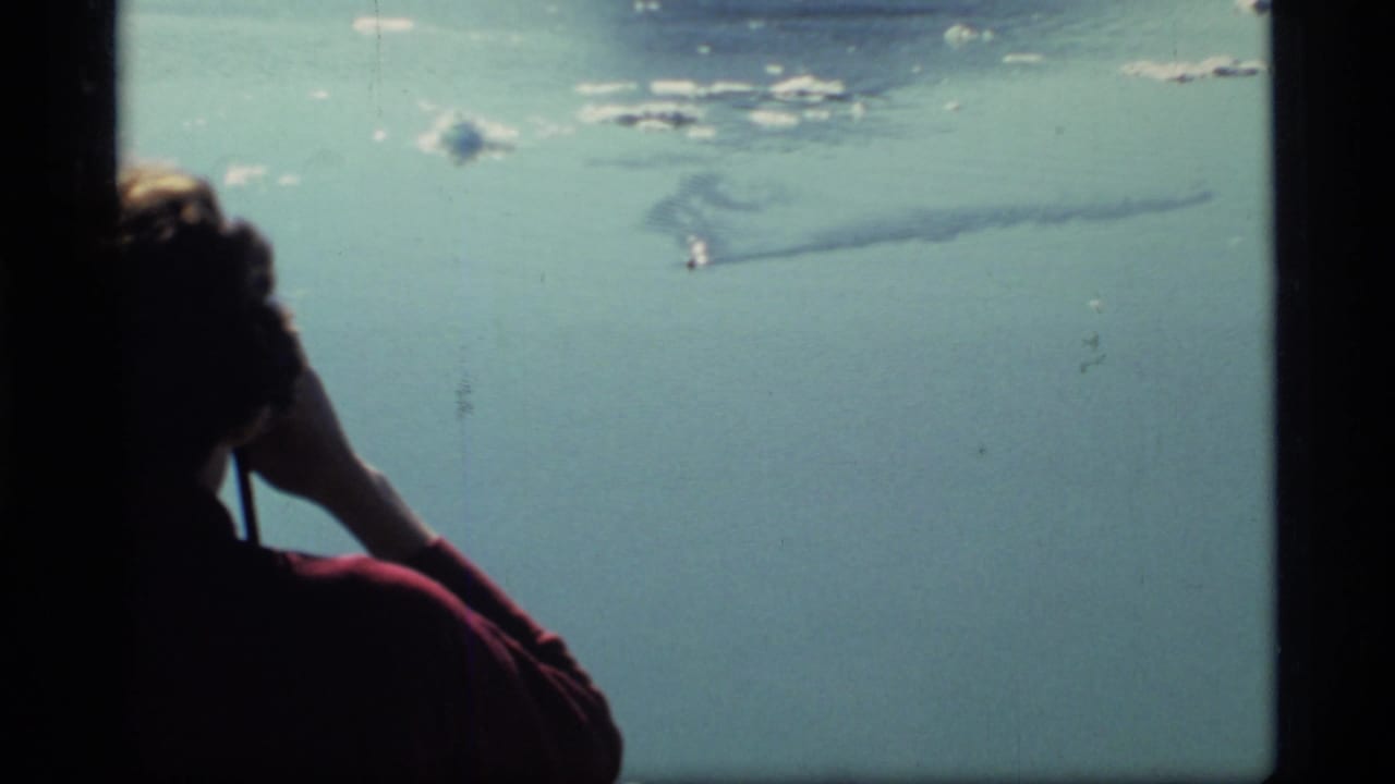 1977 Glacier Bay 8mm frame of a person in a red shirt watching calm water as a small boat leaves a wake offshore.