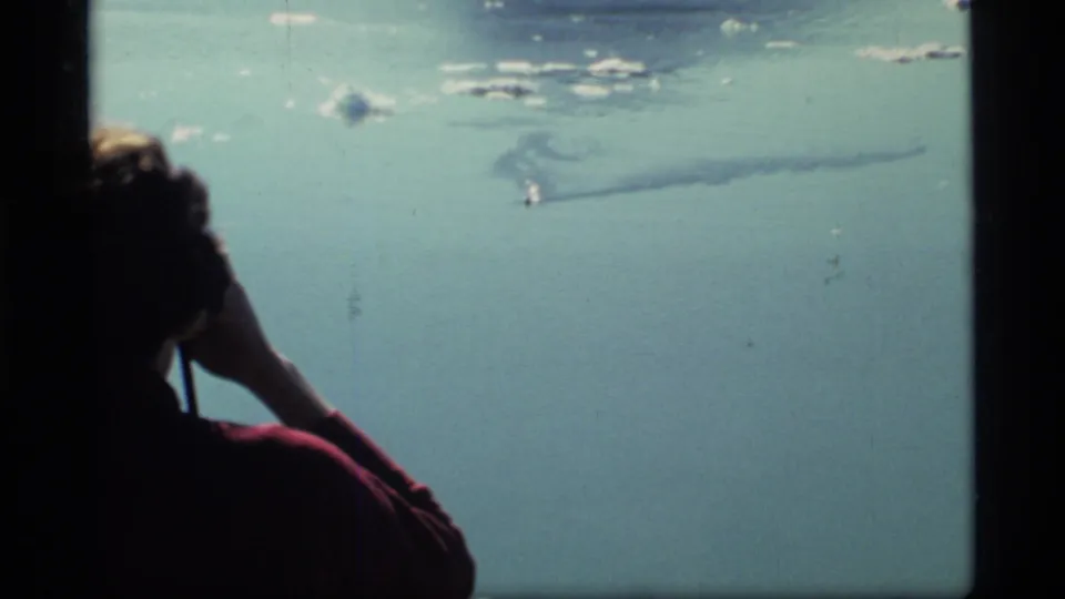1977 Glacier Bay 8mm frame of a person in a red shirt watching calm water as a small boat leaves a wake offshore.