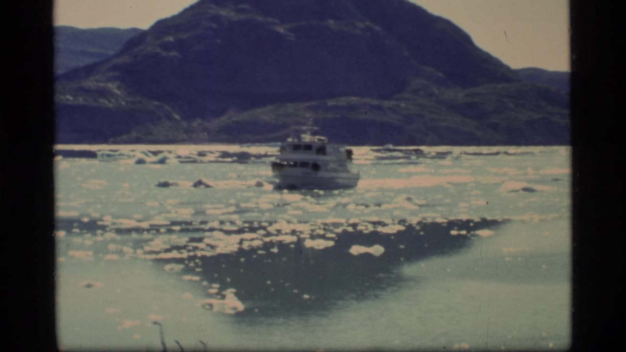 1977 8mm Glacier Bay view of a tour boat moving through pale icy water, with scattered floes and dark mountain slopes in the background.