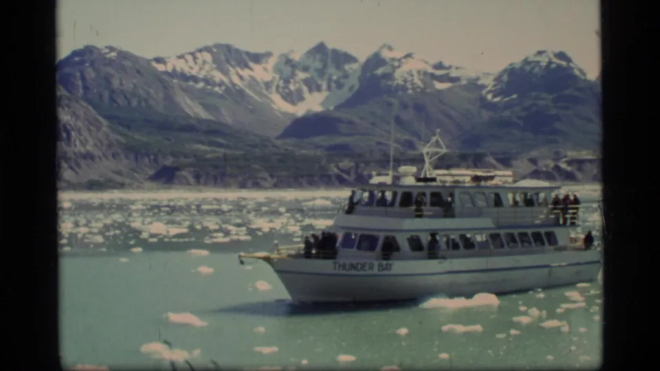 A 1977 8mm frame shows the Thunder Bay tour boat broadside among scattered ice in Glacier Bay, with passengers aboard and jagged mountain peaks beyond.