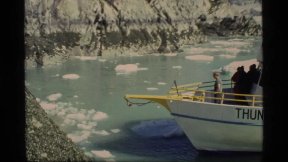 Close 1977 Glacier Bay home-movie view of the Thunder Bay bow and deck rail with passengers nearby, floating ice, and a rocky glacial shoreline behind.