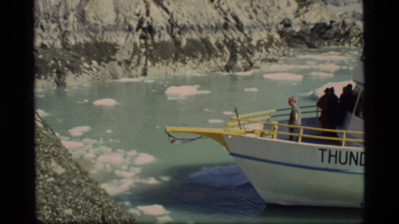 1977 Glacier Bay home-movie frame showing the Thunder boat near ice floes, with passengers on deck and rocky shoreline behind.