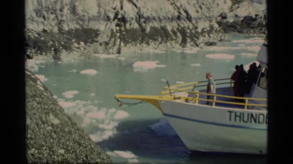 Glacier Bay 1977 frame with the Thunder boat partly cropped, passengers on deck, yellow railings, ice floes, and rugged shoreline.