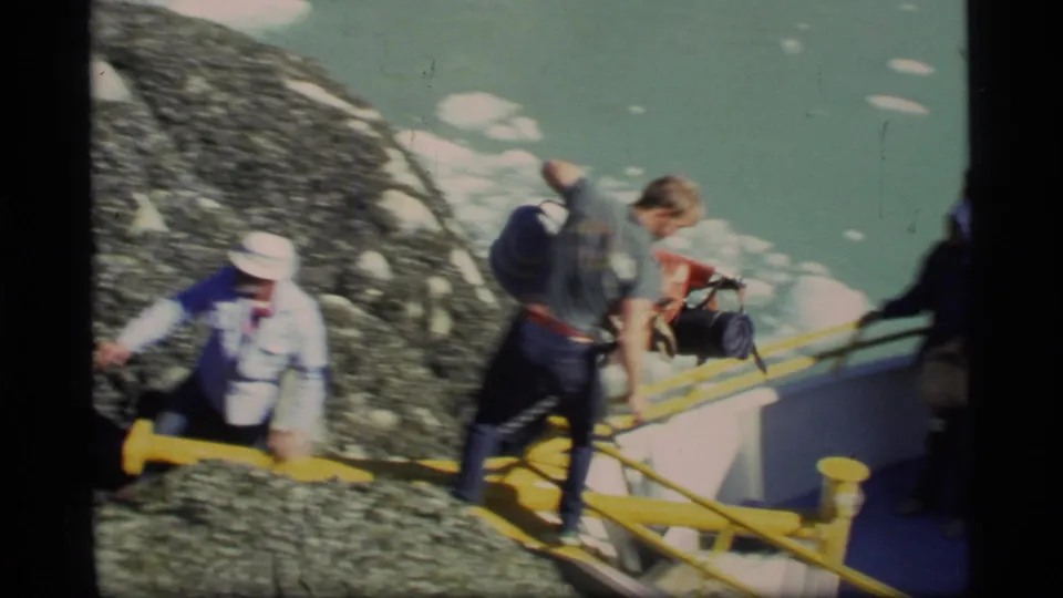 Glacier Bay 1977 frame showing passengers or workers beside the yellow-railed boat, handling supplies near ice-flecked water.