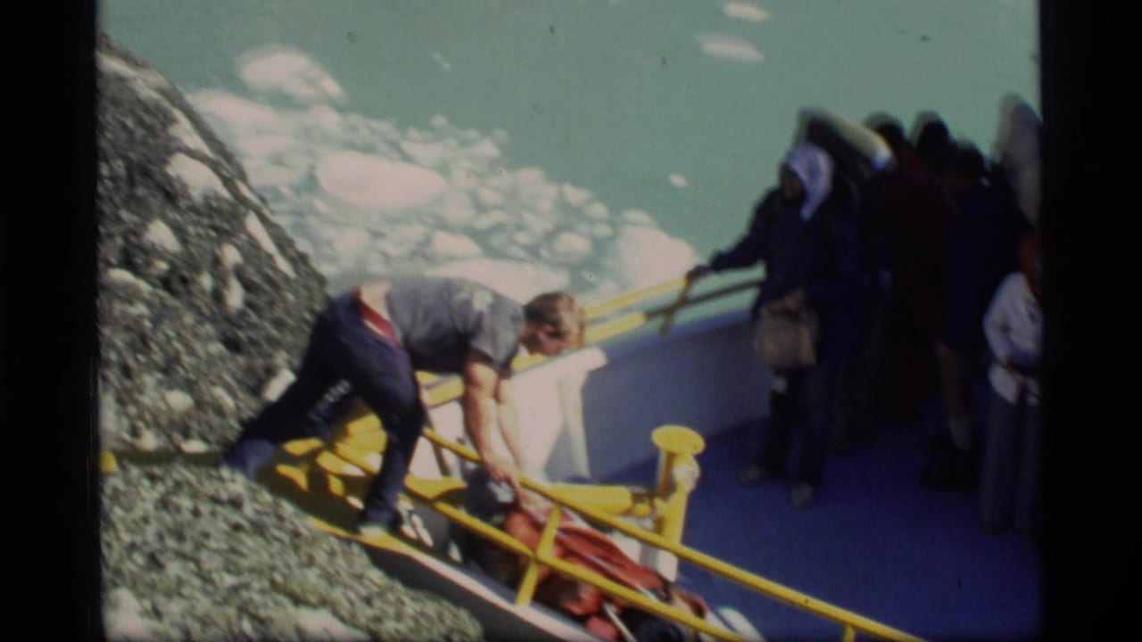 1977 Glacier Bay home-movie frame showing travelers handling gear beside a yellow-railed boat on a rocky shore.