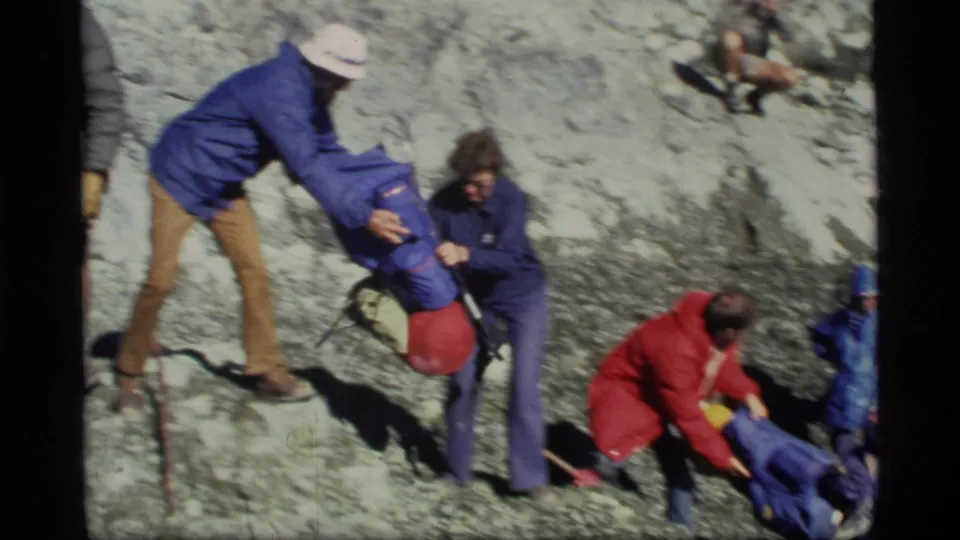 Travelers in colorful jackets appear to help each other across rocky Glacier Bay shoreline terrain.