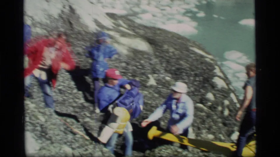 1977 Alaska expedition scene with people moving gear over uneven shoreline toward boat access.