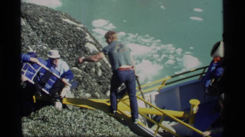 Glacier Bay home-movie frame showing people boarding or loading a yellow-railed boat from rocky shore.