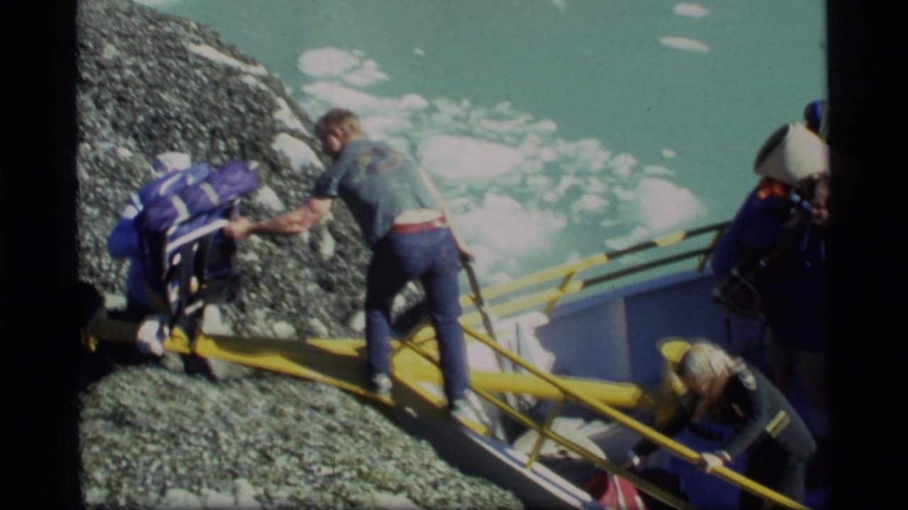 1977 Glacier Bay home-movie frame showing adults carefully boarding a small boat from gray rock beside floating ice.