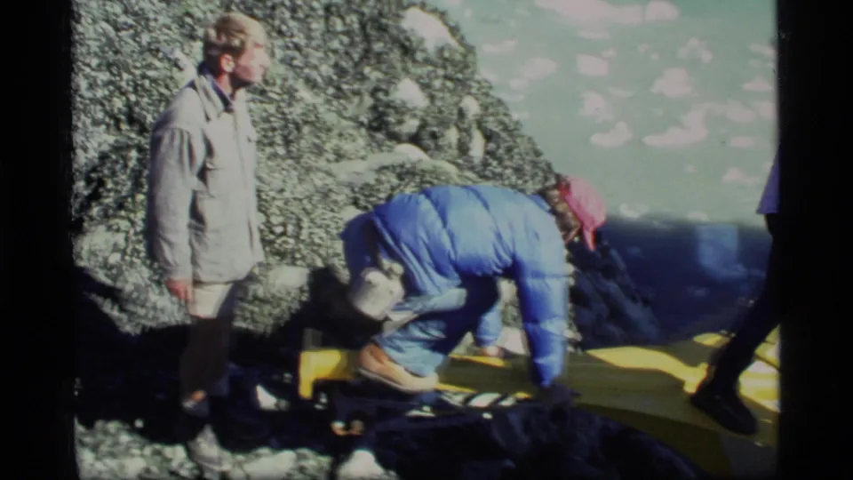 In Glacier Bay, an adult in a blue jacket and red cap crouches on a yellow boat frame while another person stands nearby.
