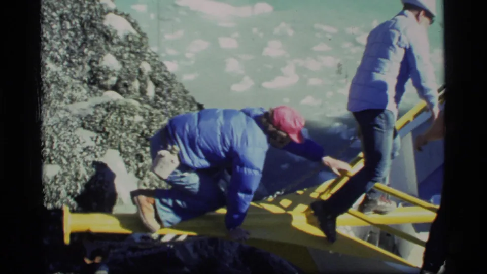 A Glacier Bay traveler in a blue jacket reaches toward a yellow rail while boarding from a rocky shoreline in 1977 footage.