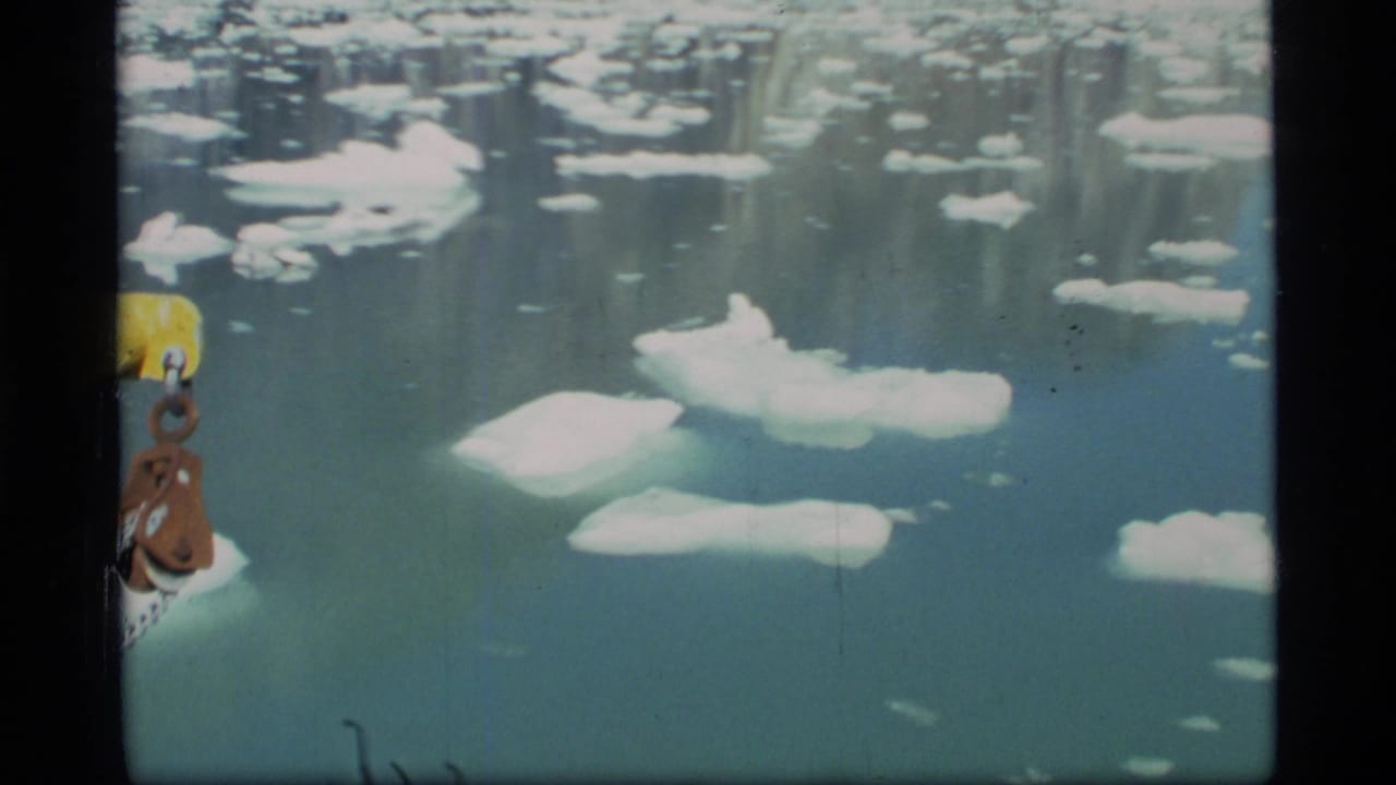 1977 8mm Glacier Bay view over icy water, with scattered white floes and a yellow-brown ship component at the left edge.