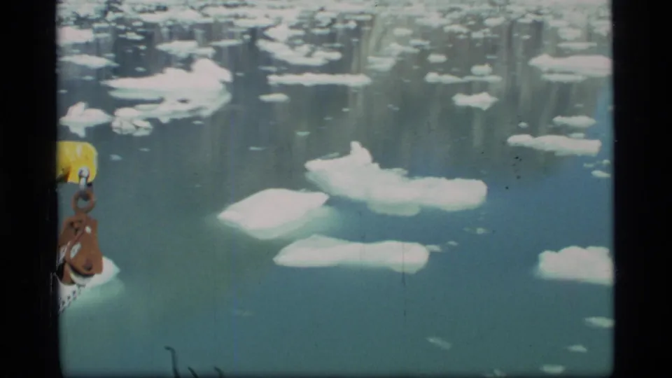 1977 8mm Glacier Bay view over icy water, with scattered white floes and a yellow-brown ship component at the left edge.