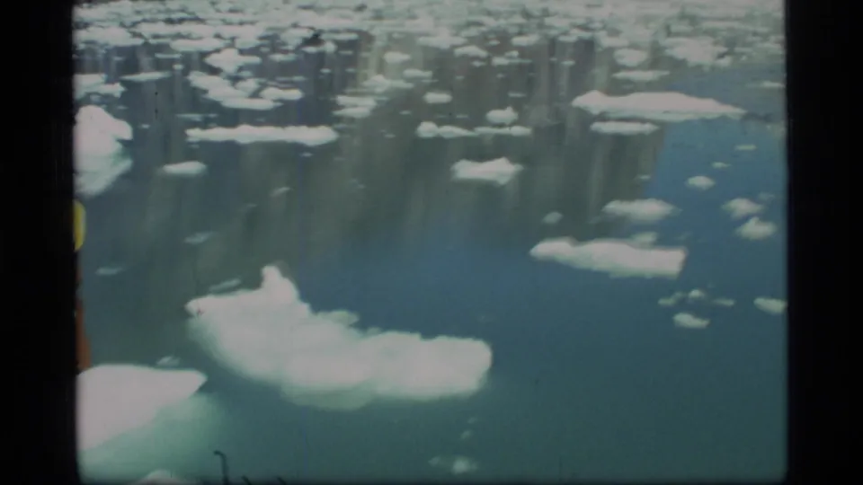 1977 Glacier Bay shipboard frame showing a larger ice floe ahead amid scattered ice on calm blue-green water.