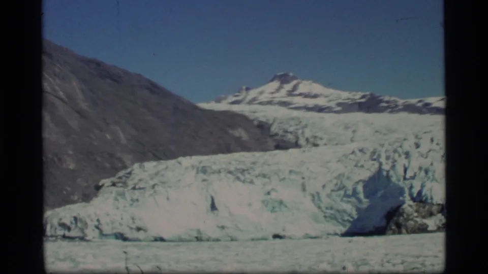 Glacier Bay landscape frame showing pale glacier ice, water, dark slopes, and snow-capped mountains beneath clear blue sky.