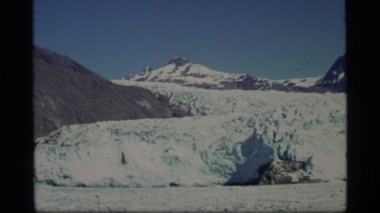 Glacier face below snow-covered mountains and blue sky, framed by dark film borders.
