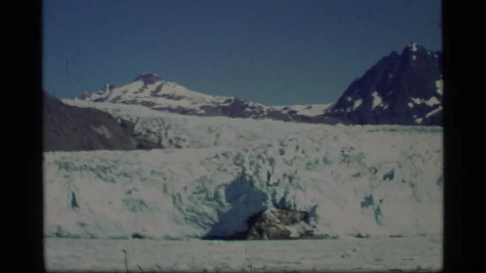 1977 Glacier Bay home-movie frame with a broad glacier face, blue ice texture, distant snow ridge, and dark mountain peak.