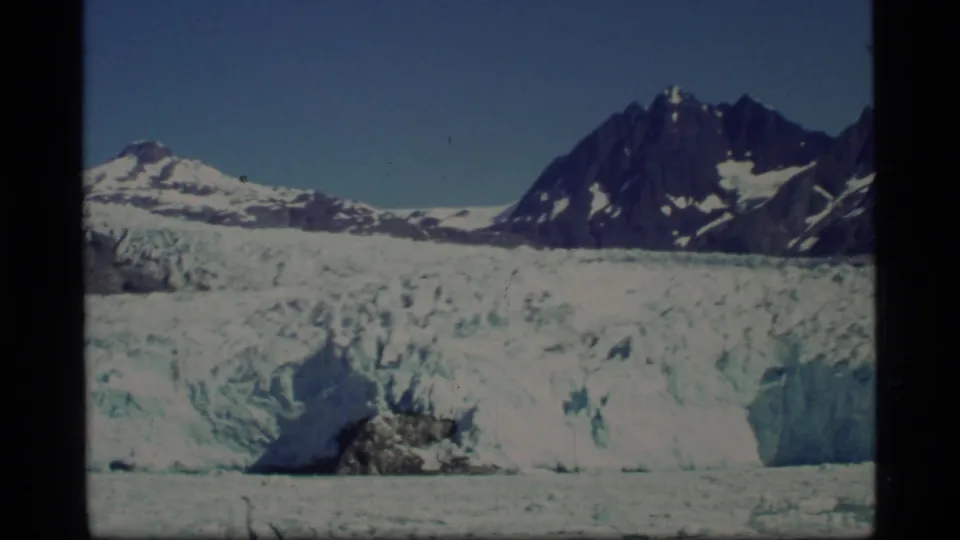 1977 8mm frame of Glacier Bay with textured ice, a low rocky outcrop, snowfields, and steep dark mountains.