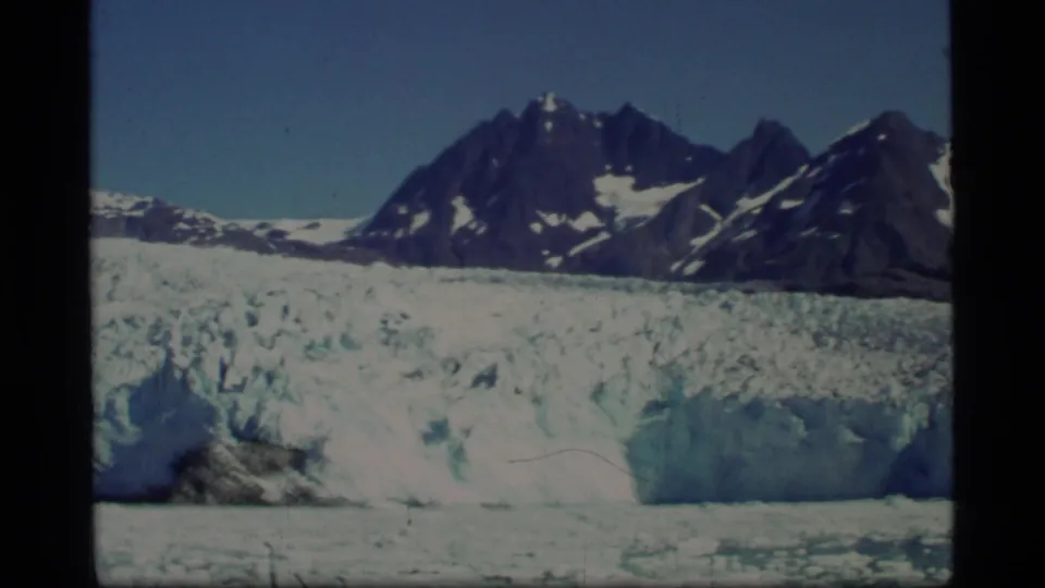 1977 Glacier Bay frame showing a glacier edge, blue ice mass, jagged mountain peaks, snow patches, and clear sky.