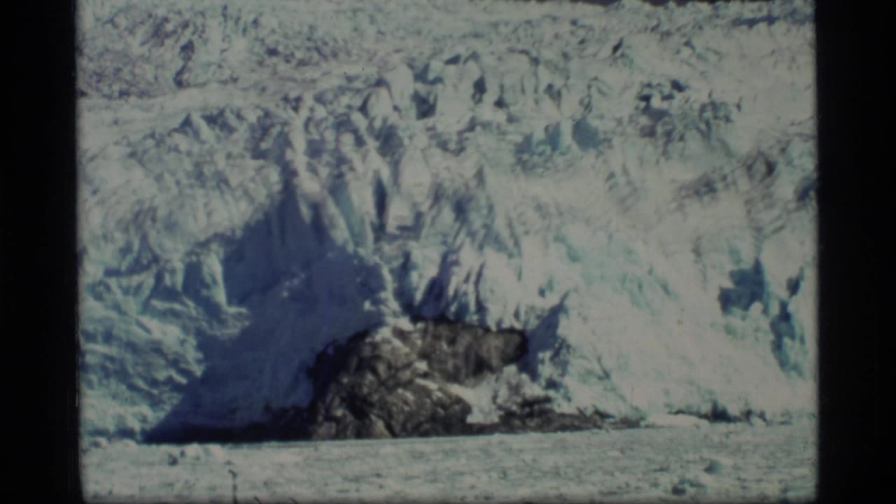 1977 8mm Glacier Bay view of a blue-white glacier face with exposed dark rock and icy water below.