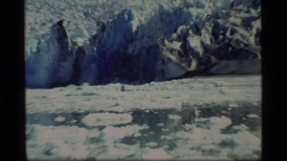 1977 Glacier Bay home-movie frame with floating ice, calm water, and a blue glacial wall in the distance.