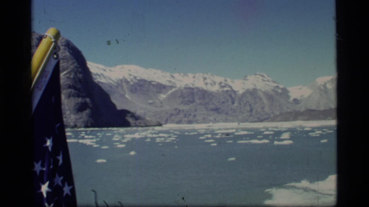 American flag beside icy bay water with snow-covered mountains in the distance.