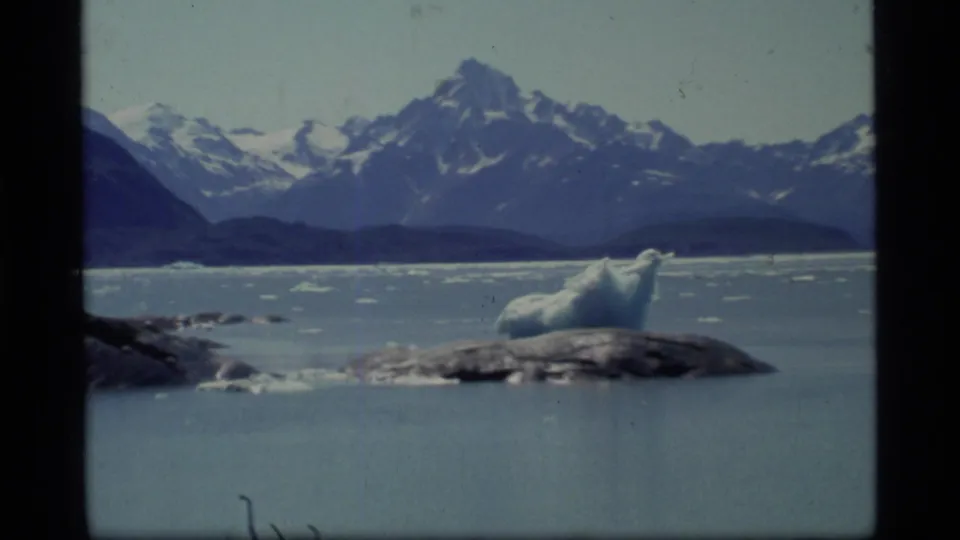 Glacier Bay boat-view frame showing a large ice chunk near rocks, calm icy water, and dark snow-streaked mountains in the background.