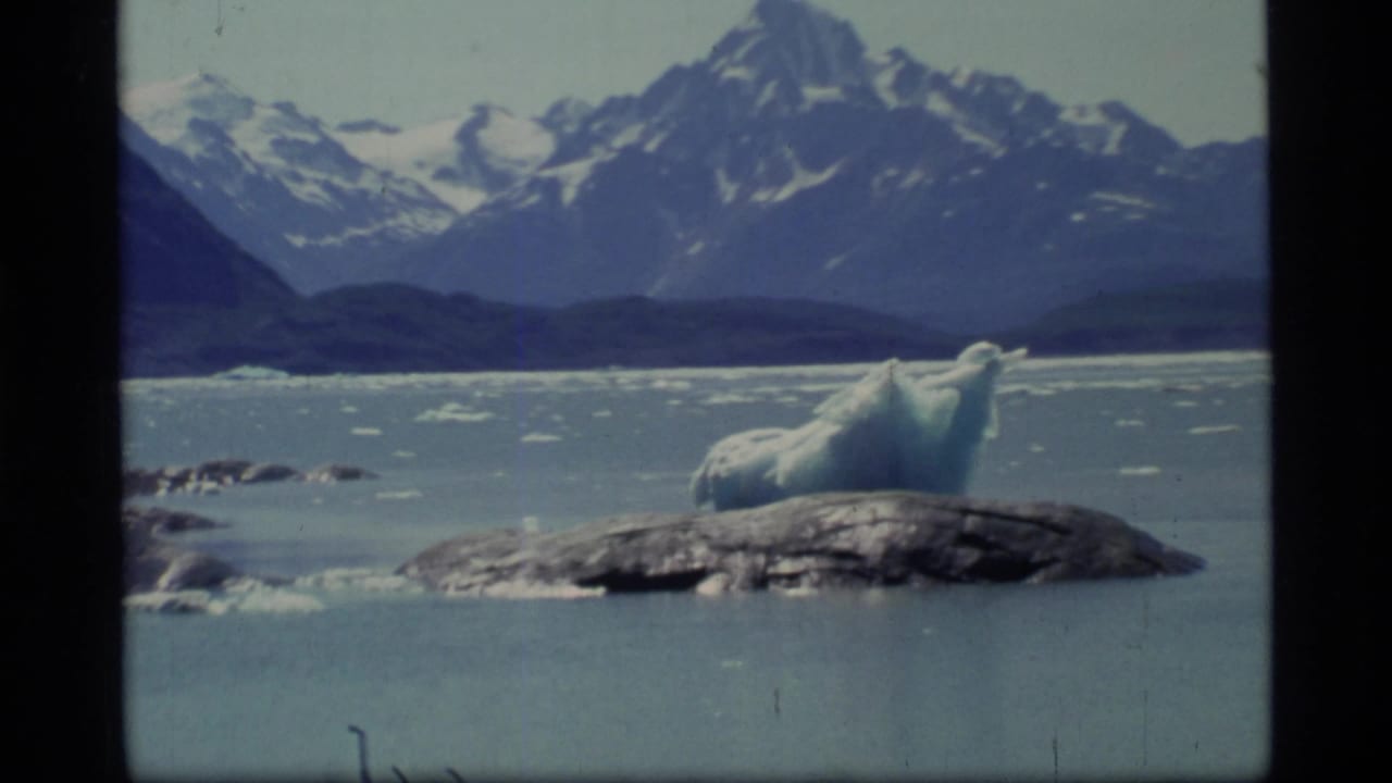 Blue-white iceberg beside a rocky outcrop in icy water with mountains behind it.