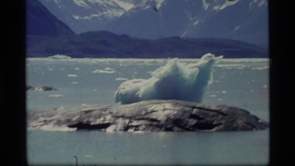 1977 Glacier Bay frame with a large blue-white iceberg, scattered ice on the water, dark shoreline, and snow-covered mountains beyond.