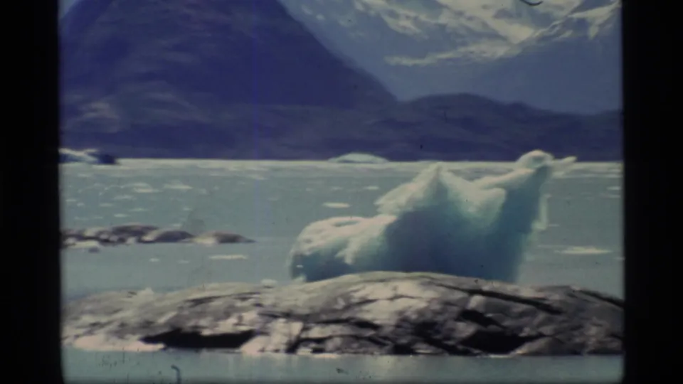 Closer 1977 Glacier Bay 8mm view of a blue-white iceberg rising from pale water near a dark rock ledge and mountain backdrop.
