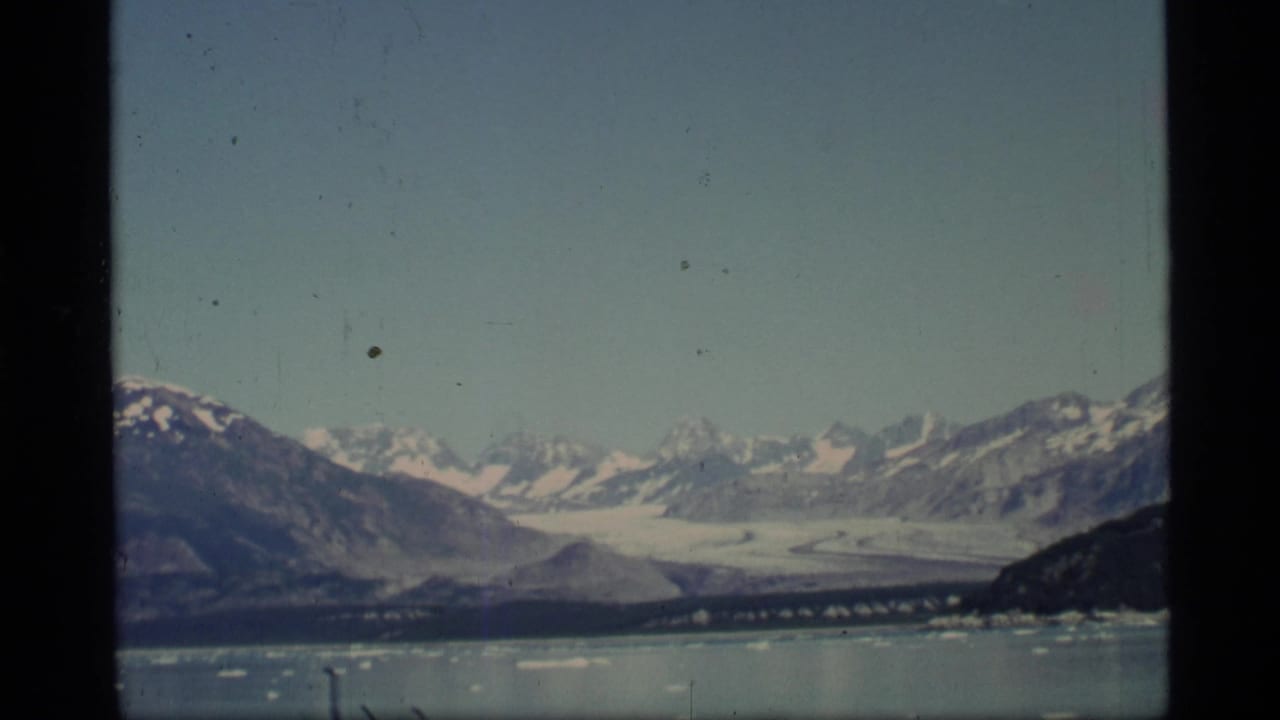 1977 8mm home-movie view of Glacier Bay with a broad glacier below snow-capped mountains and scattered ice on the water.