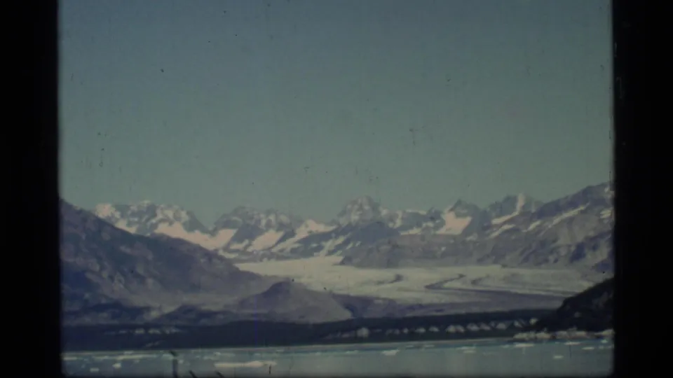 Glacier Bay, Alaska, seen on 8mm film with a wide glacier, distant peaks, calm water, and small pieces of ice near shore.