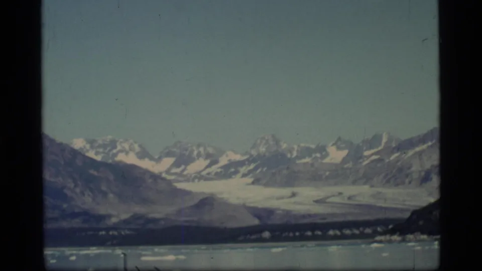 8mm frame from Glacier Bay showing a glacier valley beneath snow-covered peaks, with water and small ice pieces in the foreground.