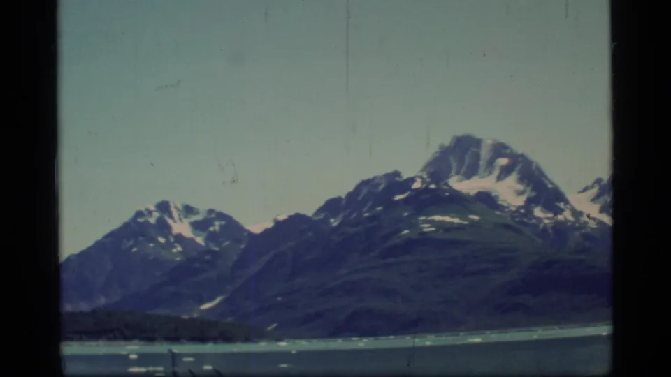 1977 Glacier Bay home-movie frame showing darker mountain slopes, snow patches, calm water, and scattered ice near the shoreline.
