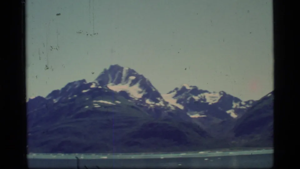 Glacier Bay landscape frame with layered mountain ridges, snow patches, pale sky, and cold water in the foreground.