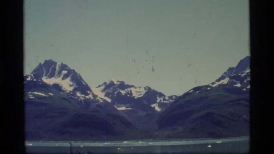 1977 home-movie frame from Glacier Bay showing a wider mountain range across water with small ice pieces near the shore or boat.