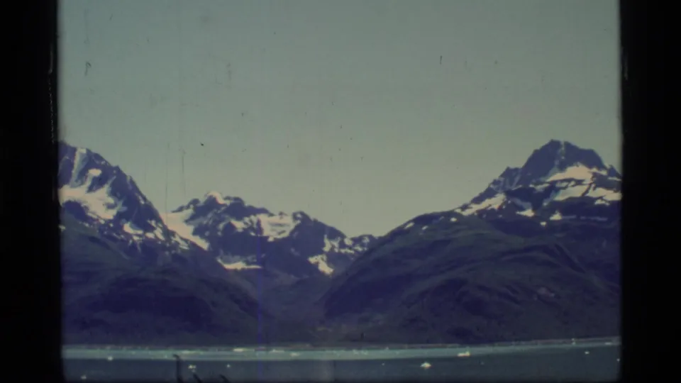 Glacier Bay 8mm frame showing snow-marked peaks, dark sloping ridges, pale sky, and scattered ice on the water.