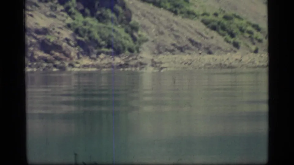 1977 8mm Glacier Bay view of still water, rocky shore, and a steep green-brown hillside with visible film wear.